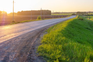 Gün batımında kıvrımlı bir yolda yol kenarında yeşil çimenler ve elektrik direkleri.