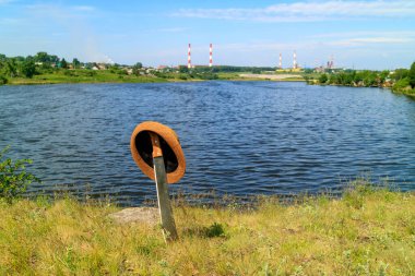 Göletin arka planına ve yere sıkışmış fabrika borularına karşı kahverengi kenarlı bir şapka takıyordu.