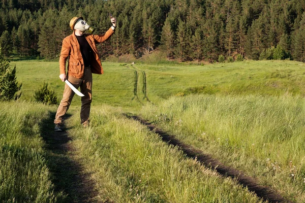 Un homme vêtu d'une veste brune et d'un jean se tient dans le champ à la main une épée sur sa tête un chapeau et un masque à gaz prend un selfie — Photo