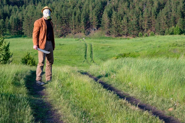 Un homme avec un chapeau et un masque à gaz dans ses mains, se tient sur la route des champs — Photo