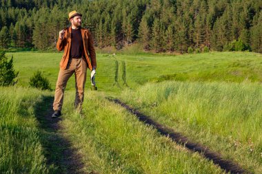 Kafasında kahverengi elbiseli bir adam yeşil bir tarlada duruyor omzunda bir kılıçla.