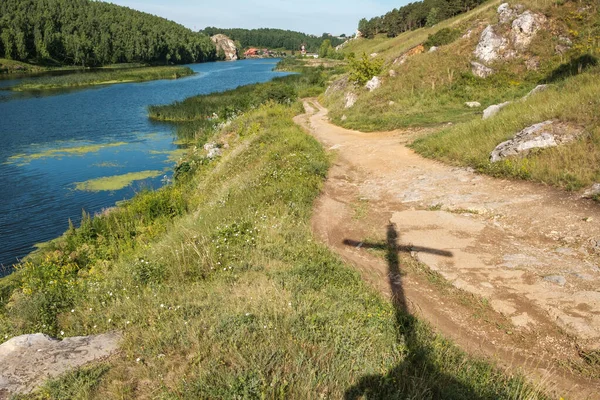 A sombra da cruz cai na estrada que corre ao longo do rio — Fotografia de Stock