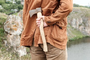 Nehir kıyısında duran bir adam arkasında bir balta saklar. Arka plandaki kayalar ve nehir