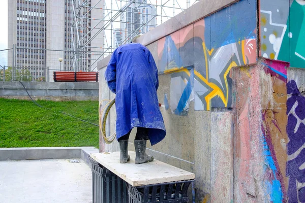 Rusya, Yekaterinburg, 14 Ağustos 2019. Yağmurluk giyen bir adam duvardaki graffitileri su ve kimyasallarla temizliyor. Modern sanatın yıkımı