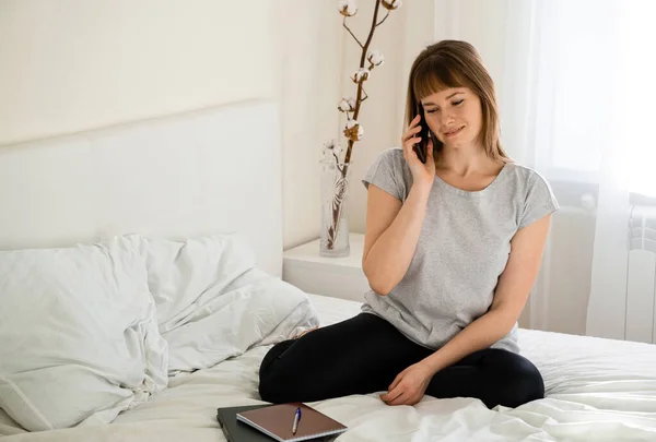 Gülümseyen genç kız yatakta oturuyor ve telefonda konuşuyor.
