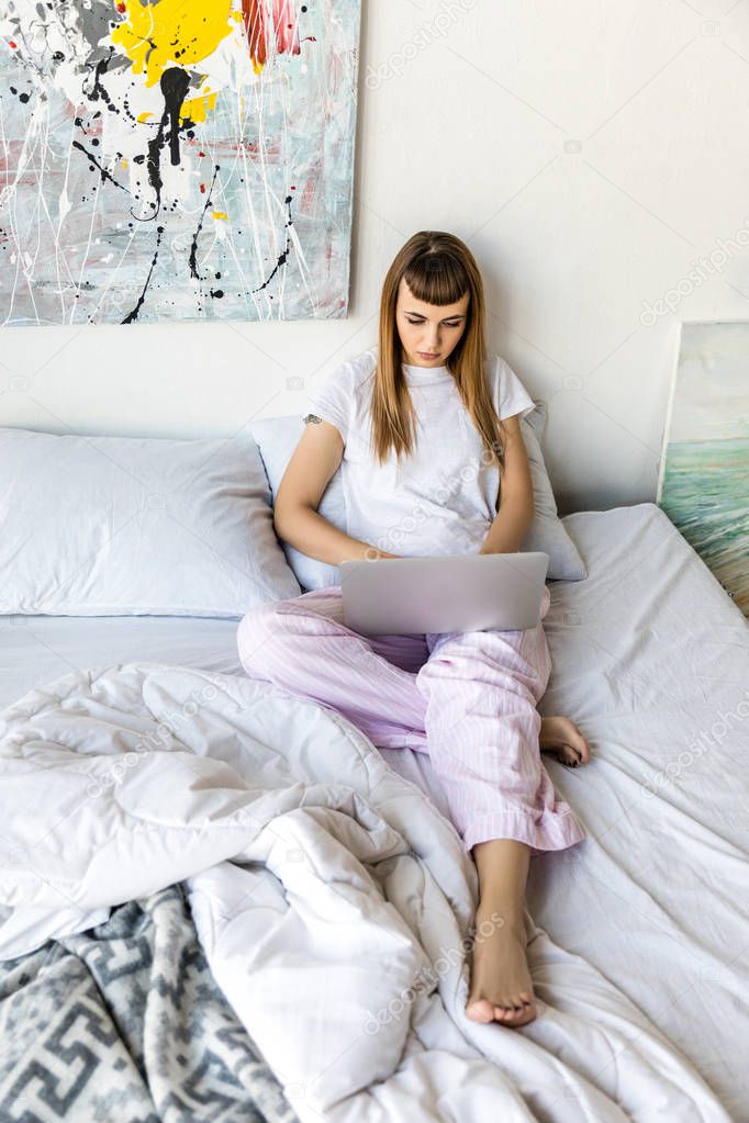 focused young woman using laptop while resting in bed in morning at home