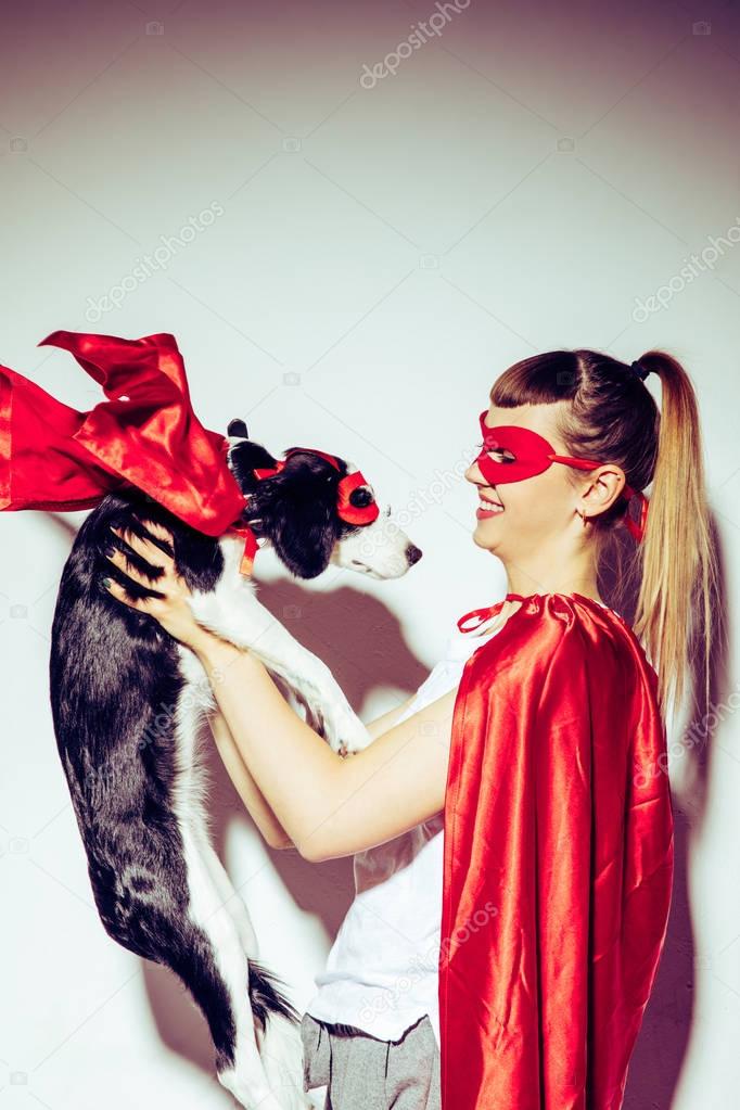side view of cheerful woman looking at little puppy in superhero costume in hands
