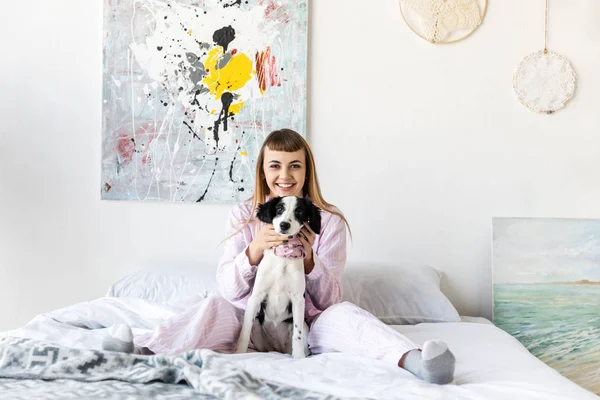 Щаслива жінка в піжамі і милий маленький щеня відпочиває на ліжку вранці вдома — стокове фото