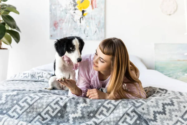 Portrait of woman holding puppys paw while lying on bed together in morning — Stock Photo