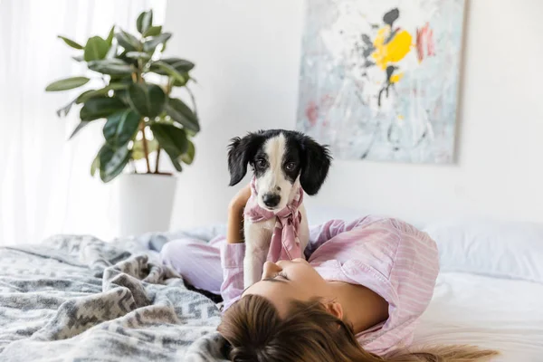 Young woman in pajamas lying on bed together with black and white puppy — Stock Photo