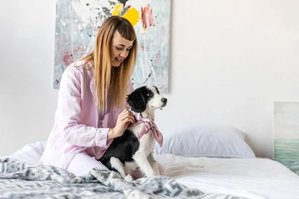 Smiling woman in pajamas and cute little puppy resting on bed in morning at home — Stock Photo