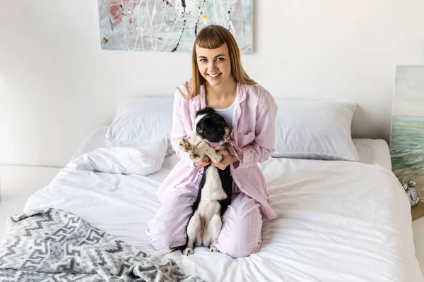 Mulher feliz em pijama e filhote de cachorro bonito descansando na cama de manhã em casa — Fotografia de Stock