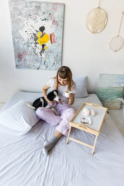 Mulher beber café enquanto descansa na cama junto com filhote de cachorro — Fotografia de Stock