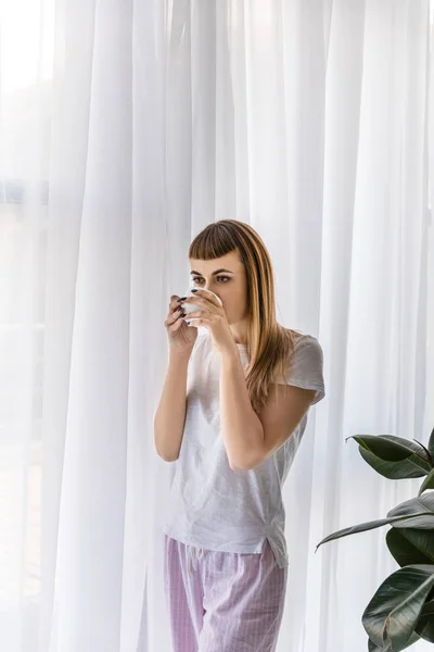 Giovane donna in piedi alla finestra e bere caffè al mattino — Foto stock