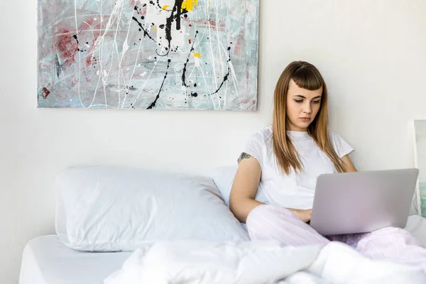 Focada jovem mulher usando laptop enquanto descansa na cama de manhã em casa — Fotografia de Stock