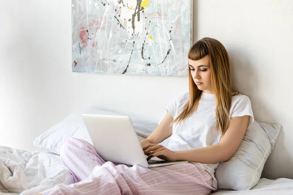 Fokussierte junge Frau benutzt Laptop, während sie morgens zu Hause im Bett liegt — Stockfoto