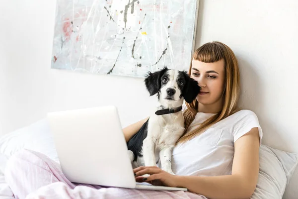 Retrato de mujer joven usando el ordenador portátil con cachorro cerca por la mañana en casa - foto de stock