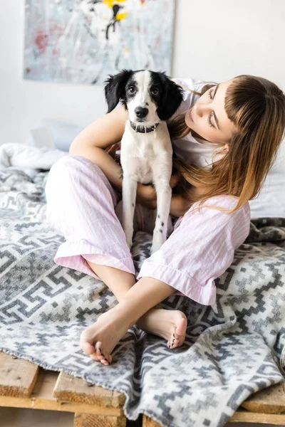 Jovem mulher abraçando filhote de cachorro enquanto sentado na cama de manhã em casa — Fotografia de Stock
