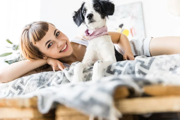 Portrait of happy woman looking at camera while resting in bed together with puppy — Stock Photo