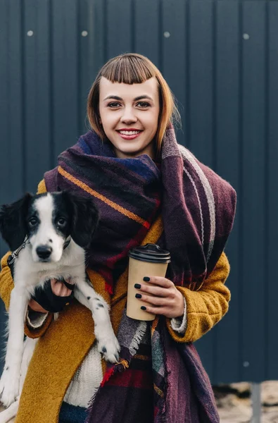 Retrato de mujer sonriente sosteniendo cachorro y café para ir en las manos mientras está de pie en la calle - foto de stock