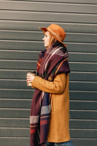 Vista lateral de mujer atractiva en chaqueta de otoño y sombrero con café para ir en las manos - foto de stock
