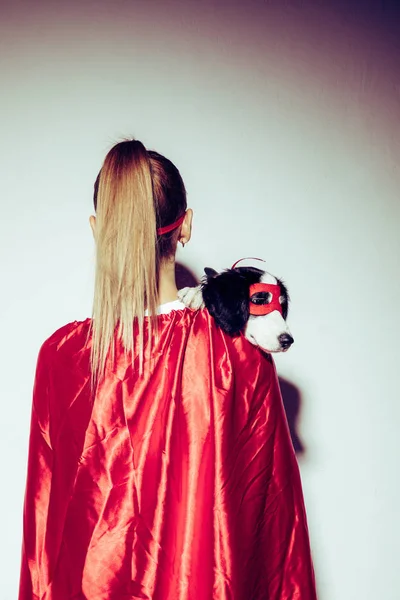 Back view of woman in red cape holding little puppy in superhero mask — Stock Photo