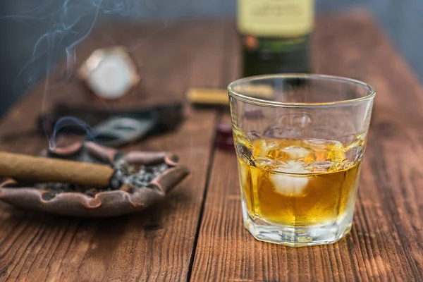 A glass of whiskey with ice on a table in the foreground and a cigar with an ashtray, a guillotine, a lighter, a fragment of a whiskey bottle and a clock in the background in defocus