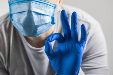 Koruyucu bir tıbbi eldiven giymiş bir el koruyucu bir maskenin içinde yüzünün yanında iyi bir jest gösteriyor.