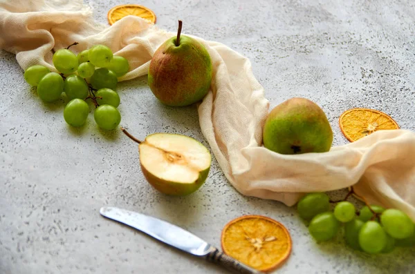 Three pears with green grapes and three dried pieces of orange decorated with silver vintage knife and light brown cloth on gray concrete background close up