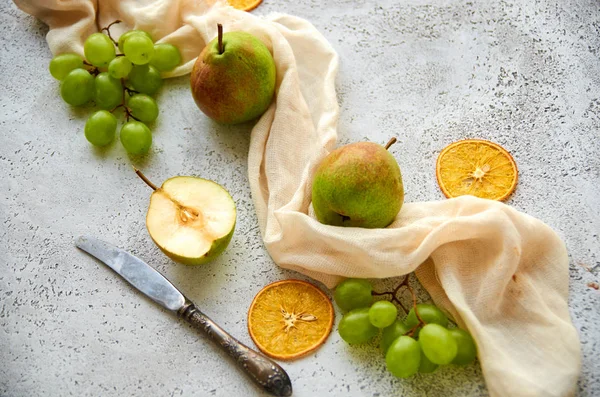 Three pears with green grapes and three dried pieces of orange decorated with silver vintage knife and light brown cloth on gray concrete background close up