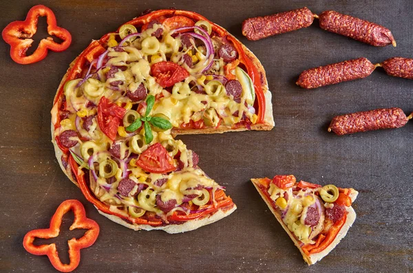 Piece of pizza isolated with salami, tomatoes, bell pepper, onion, green olives, corn, cheese and spices. Sliced pizza, little sausages salami and fresh bell pepper on brown wooden table — Stock Photo, Image