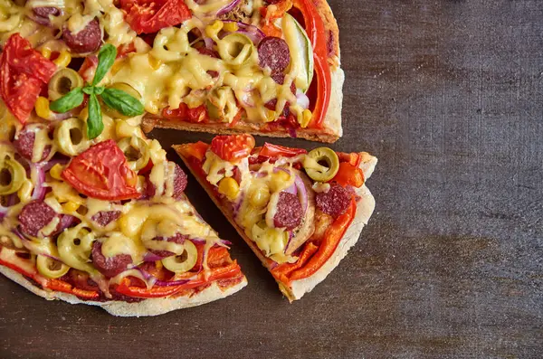 Pizza en rodajas y trozo de pizza aislado con salami, tomates, pimiento, cebolla, aceitunas verdes, maíz, queso y especias sobre fondo de madera marrón — Foto de Stock