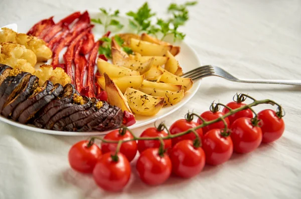 Жареный картофель, баклажаны, перец и цветная капуста на белой тарелке украшены вилкой, свежими помидорами черри и петрушкой — стоковое фото