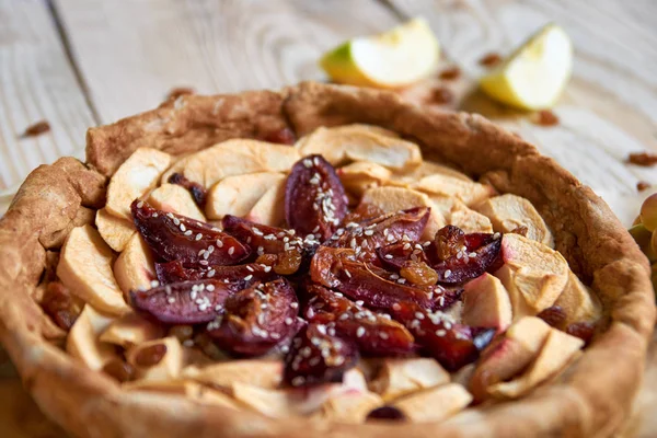 A homemade apple plums pie decorated with fresh apples, brown raisins and sesame on light wooden background