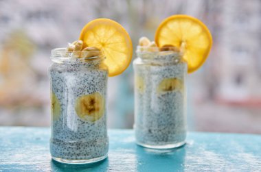 Mavi ahşap masa üzerinde iki vintage kaşık çiğ chia tohum ile iki cam kavanoz muz ile Chia puding. Chia taze meyve ile güler yüzlü. Chia puding muz ve portakal bulanık gri arka plan üzerinde