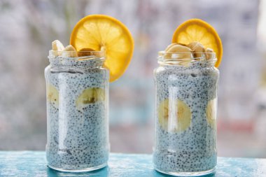 Chia puding muz mavi ahşap masa üzerinde iki cam kavanozlara ile. Chia taze meyve - muz ve portakal bulanık gri arka plan üzerinde ile güler yüzlü. Görünümü kapatın