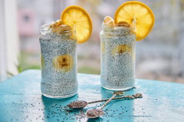 Mavi ahşap masa üzerinde iki vintage kaşık çiğ chia tohum ile iki cam kavanoz muz ile Chia puding. Chia bulanık gri arka plan üzerinde taze turuncu ile güler yüzlü. Yatay fotoğraf