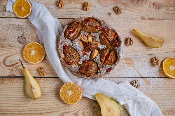 A powdered pear pie decorated with white cloth, fresh sliced pears, dried oranges, walnuts, anise stars, silver spoon on the wooden board. Healthy homemade pear pie with fresh red pomegranate seeds.