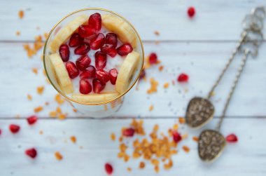 Yoğurtlu taze dilimlenmiş muz ve nar tohumları Beyaz ahşap masa üzerinde. Vintage kaşık chia, dağınık granola tahıl ve nar taneleri üzerinde arka plan bulanık. Üstten Görünüm
