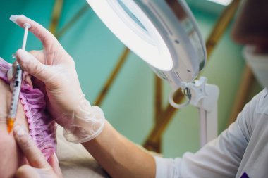 Doktor kozmetik uzmanı güzellik salonundaki genç ve güzel bir kadının cildindeki kırışıklıkları sıkmak ve düzleştirmek için yüz enjeksiyonlarını yenileme prosedürünü uyguluyor..