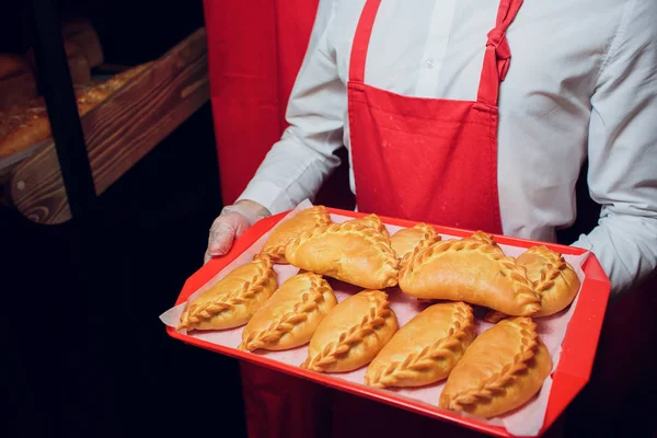 Baker mantiene una bandeja con el hombre pan fresco en panadero uniforme rojo y blanco sosteniendo una bandeja con la fabricación de pan — Foto de Stock