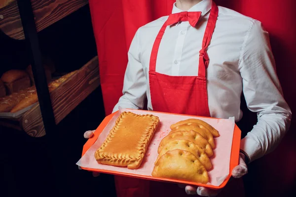 Baker mantém uma bandeja com homem de pão fresco em padeiro uniforme vermelho e branco segurando uma bandeja com panificação — Fotografia de Stock