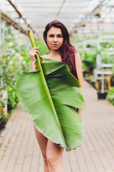 큰 바나나 잎 아래서서 카메라를 보고 있는 갈색 머리를 한 젊은 여자의 사진. — 스톡 사진