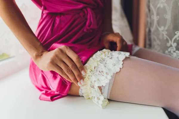 's Ochtends draagt de bruid kousen en een witte trouwjurk een kousenband op haar been, de bruid houdt haar handen vast voor de kousenband.. — Stockfoto