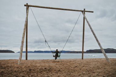 Single young Chinese woman sitting on a huge yellow swing on beach by Arctic ocean, in Teriberka, Russia. clipart