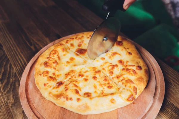 Pizza cortador de ruedas rebanando pizza de jamón con pimiento y aceitunas sobre tabla de madera en la mesa . — Foto de Stock