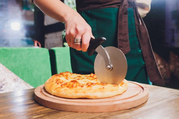 Pizza cutter wheel slicing ham pizza with capsicum and olives on wooden board on table.