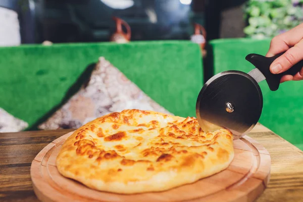 Pizza cutter wheel slicing ham pizza with capsicum and olives on wooden board on table. — Stock Photo, Image