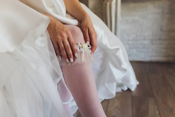 De manhã, a noiva em meias e um vestido de noiva branco usa uma liga em sua perna, a noiva está segurando suas mãos para a liga . — Fotografia de Stock