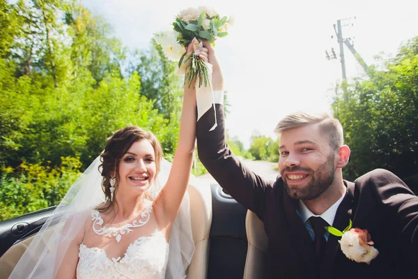 Primer plano retrato joven hermosa novia elegante pareja en un vestido blanco con un ramo de flores en el pelo y y el novio en el coche en el camino . —  Fotos de Stock